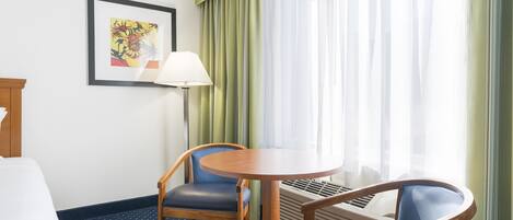 A hotel room with a bed, a desk with a lamp, two chairs, a round table, and a window with curtains.