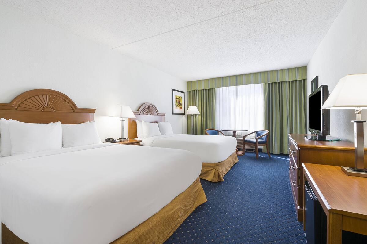 Quarto de hotel com duas camas, uma escrivaninha, uma cadeira, televisão e janela com cortinas.