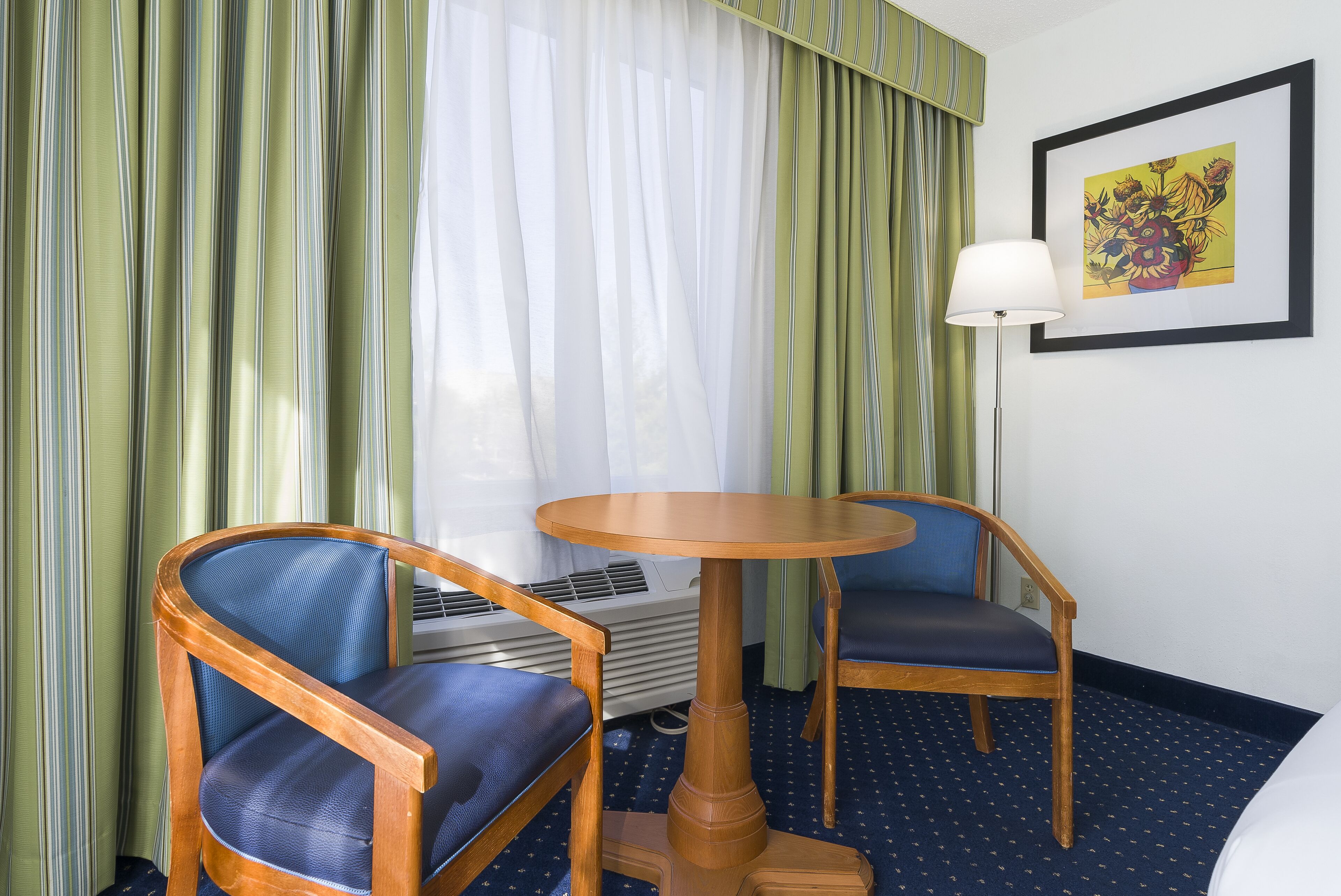 A hotel room with two wooden chairs, a small round table, a lamp, and a framed picture on the wall.