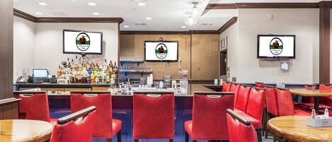 A restaurant with red chairs and tables, a bar area with a counter and various bottles, and two screens displaying the same logo.