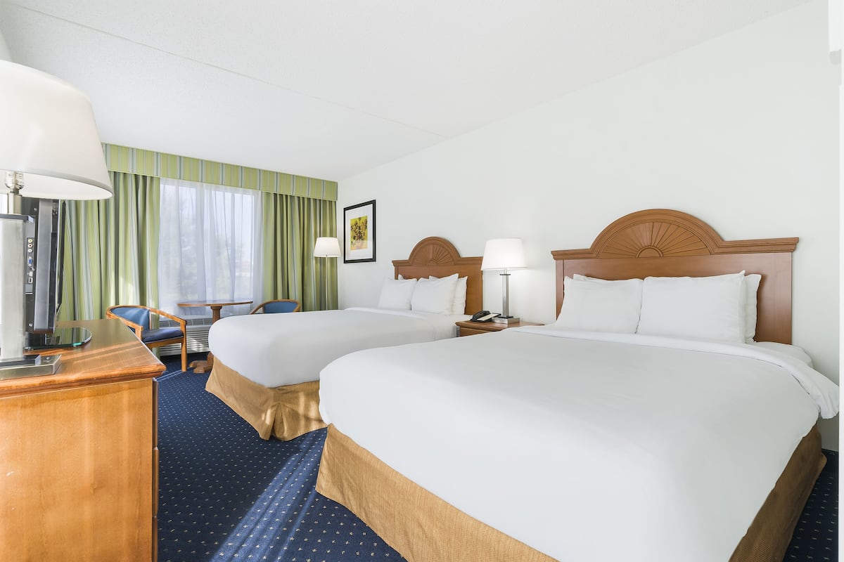 Quarto de hotel com duas camas, uma mesa, uma cadeira, um abajur e uma janela com cortinas.