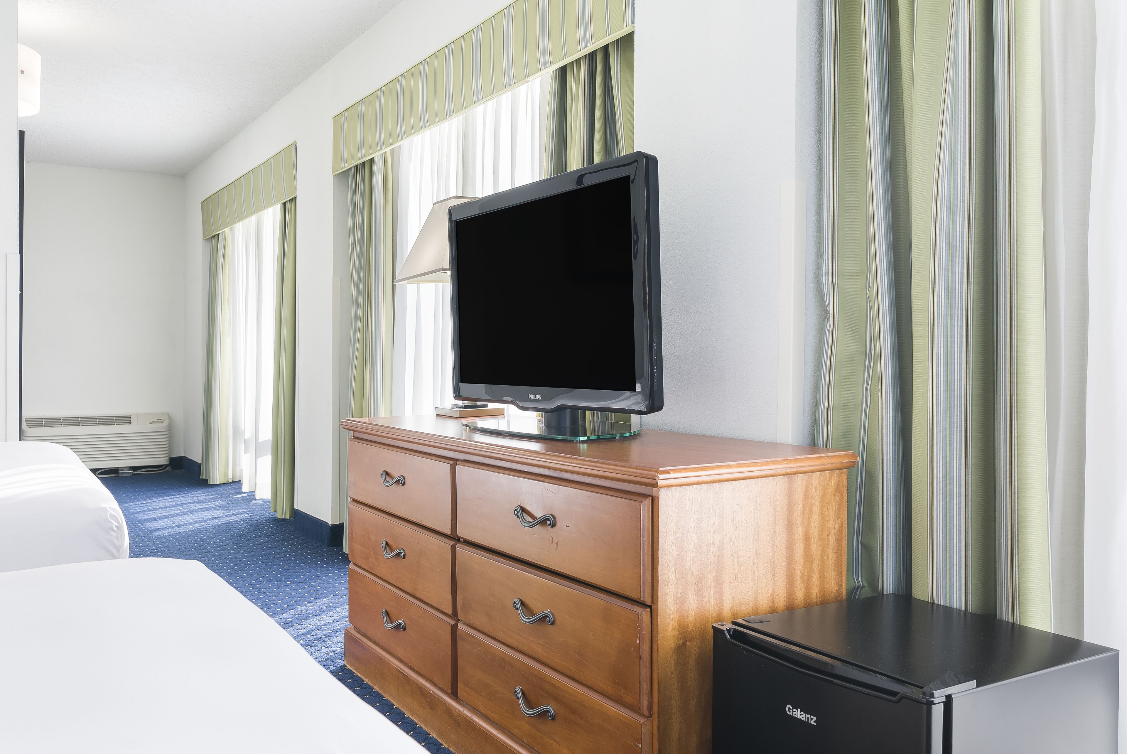 Une chambre d’hôtel avec un lit, une commode en bois avec des tiroirs, un téléviseur posé sur le dessus, un réfrigérateur noir et une fenêtre avec des rideaux à rayures.