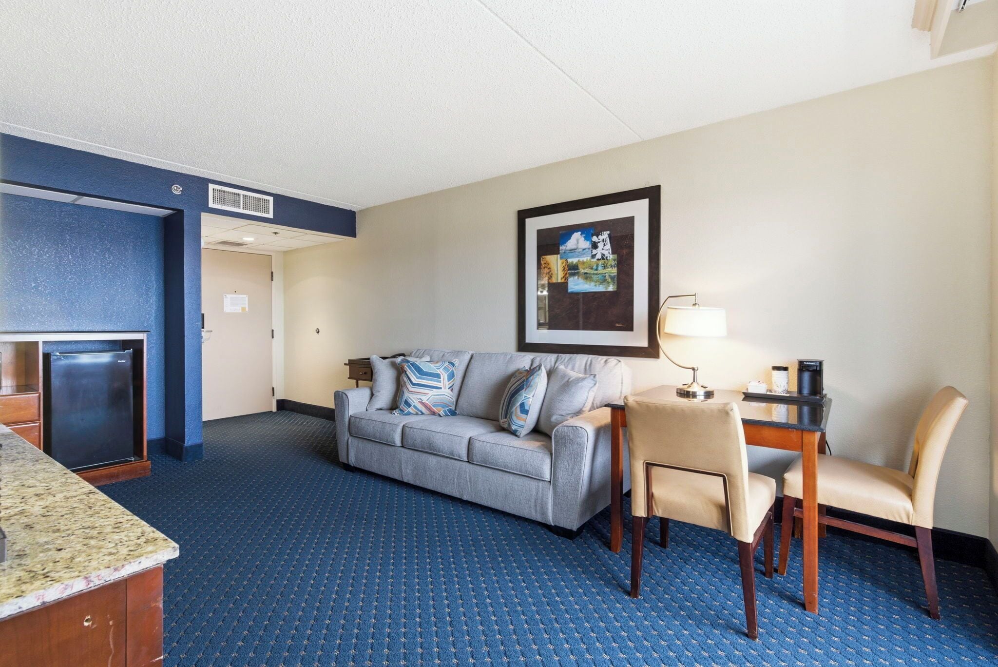 Une chambre d’hôtel avec un tapis bleu, un canapé gris, une petite table avec une lampe, une chaise et un réfrigérateur.