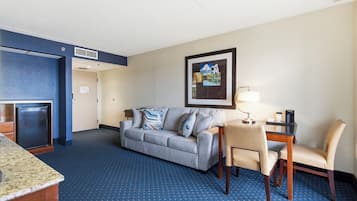 Une chambre d’hôtel avec un tapis bleu, un canapé gris, une petite table avec une lampe, une chaise et un réfrigérateur.