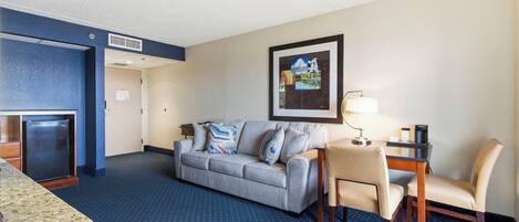 Habitación de hotel con alfombra azul, un sofá gris, una mesita con lámpara, una silla y un refrigerador.