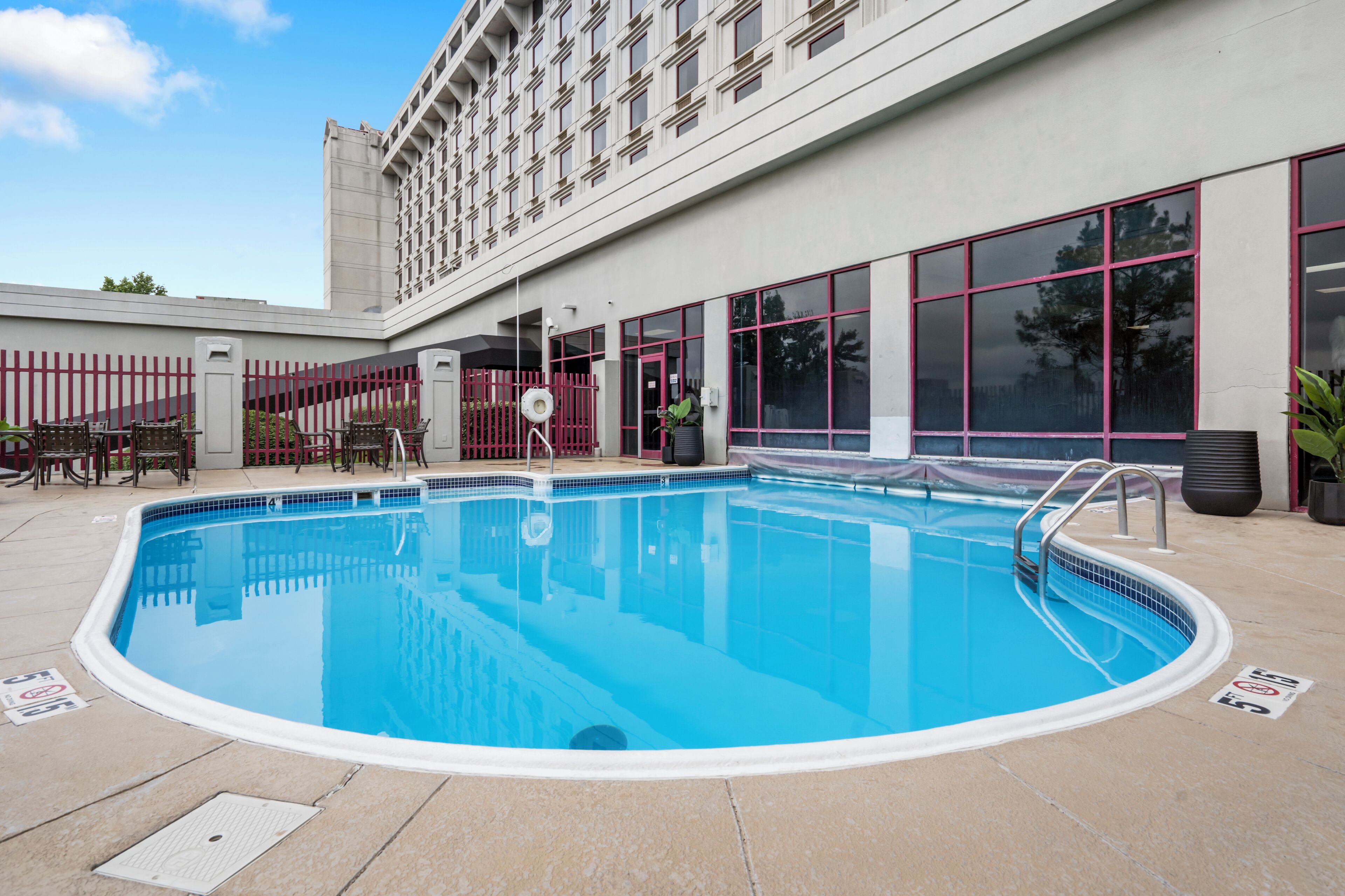 Una piscina con una cerca roja, un área de descanso cubierta y un edificio con grandes ventanales al fondo.