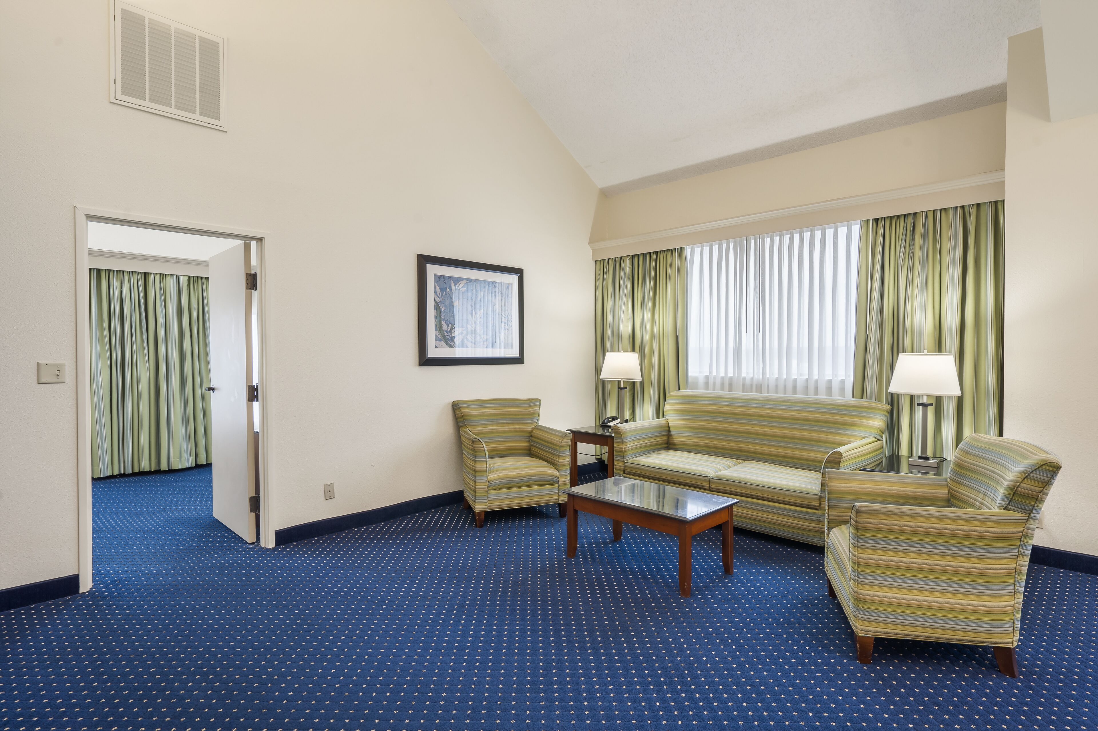 Een hotelkamer met een blauw tapijt, een bank, twee fauteuils, een salontafel en een raam met gordijnen.