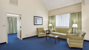 Habitación de hotel con alfombra azul, un sofá, dos sillones, una mesa de centro y una ventana con cortinas.