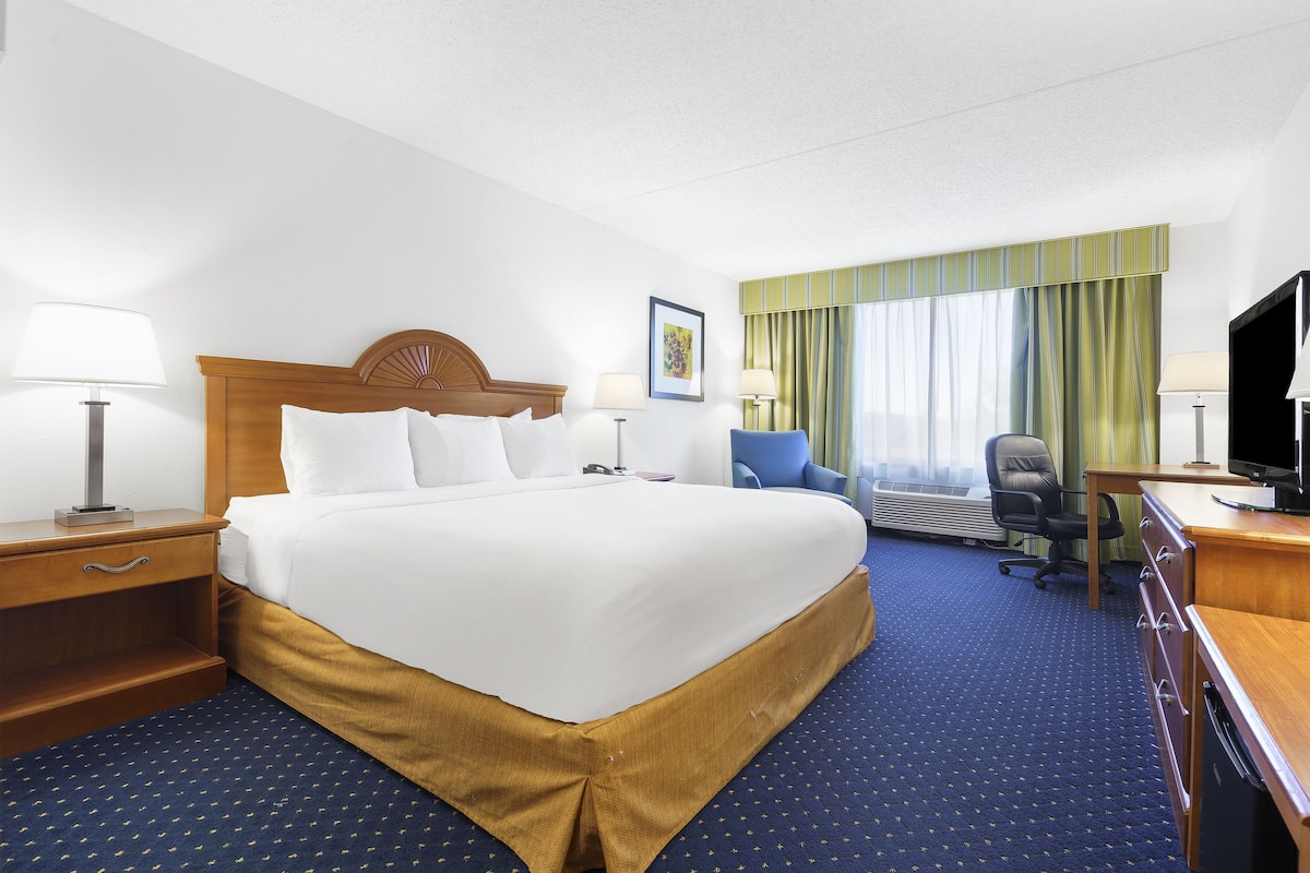 Quarto de hotel com uma cama grande, uma escrivaninha com televisão, uma cadeira, uma luminária de mesa e uma janela com cortinas.