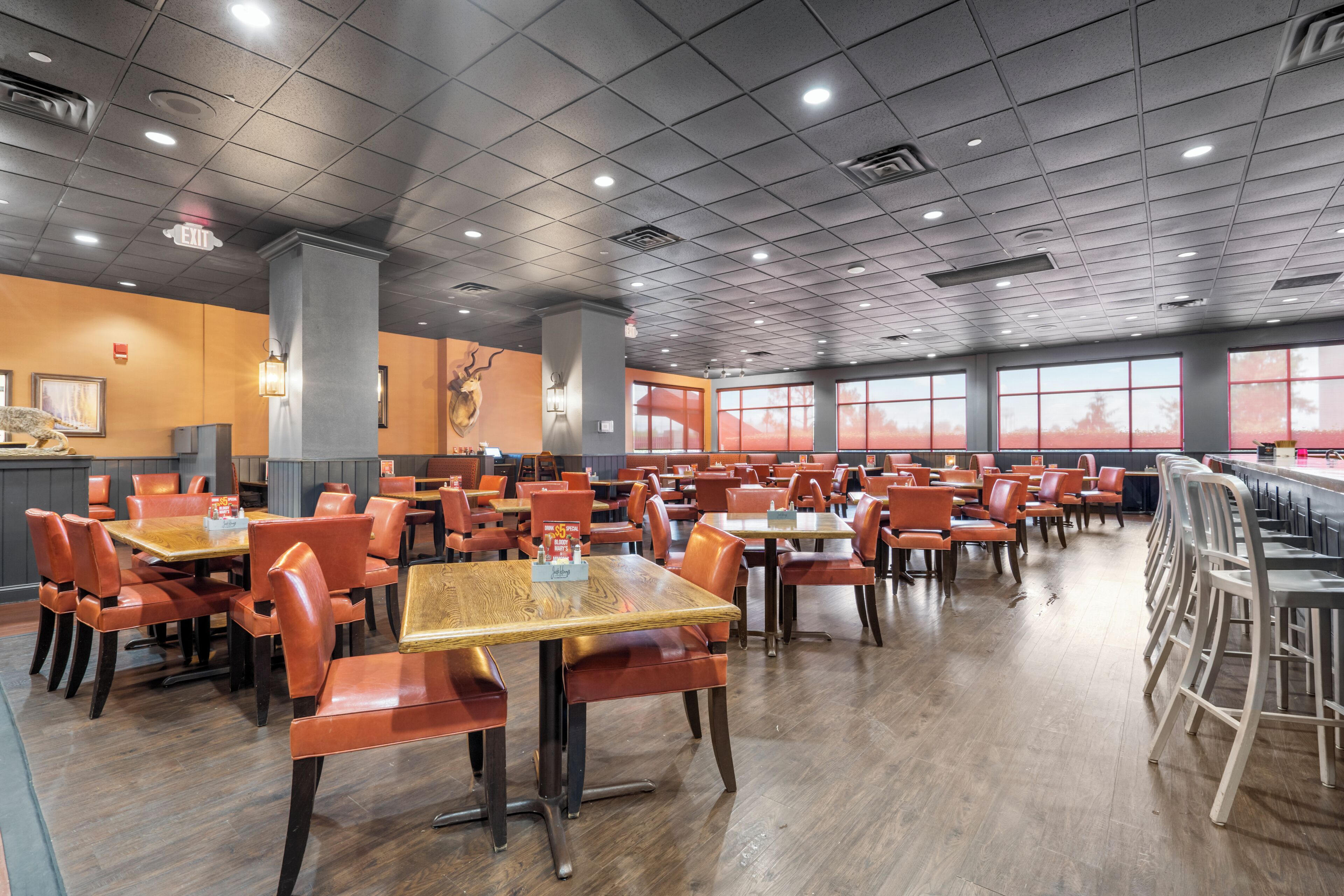 A spacious restaurant with multiple tables and chairs, a high ceiling with recessed lighting, and large windows.