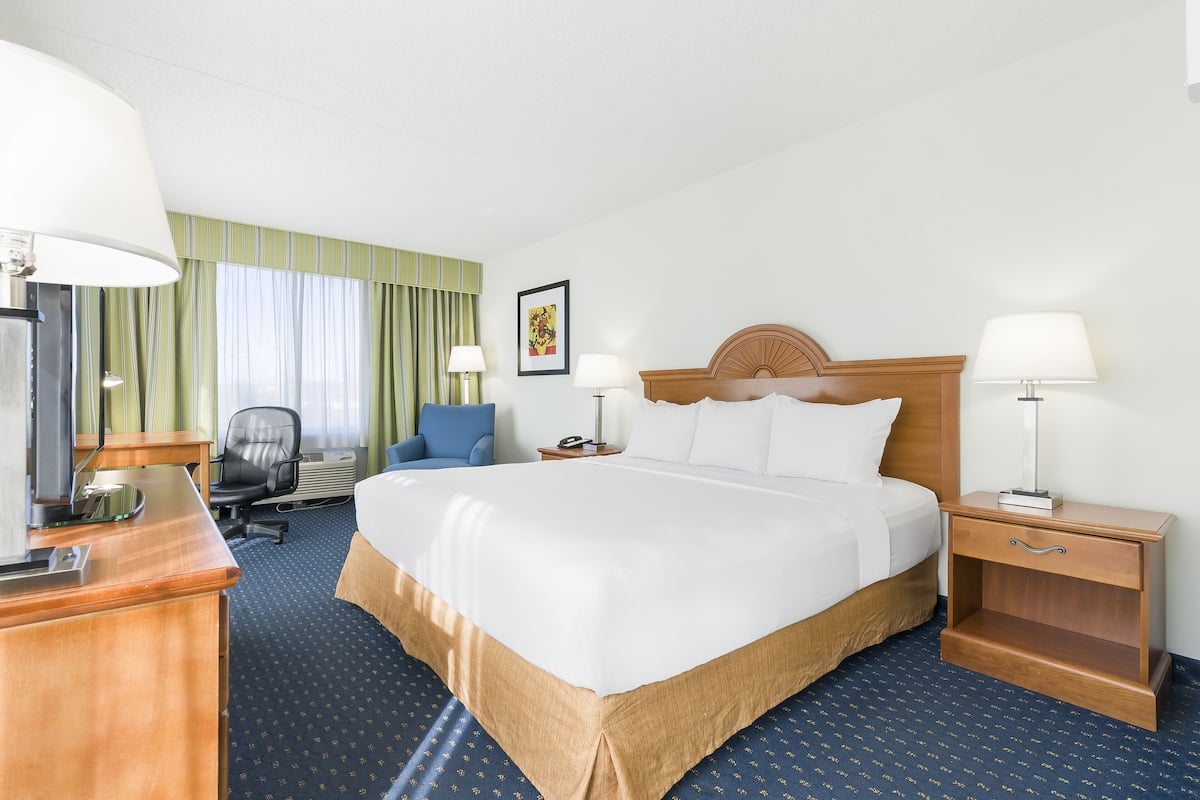 Quarto de hotel com uma cama grande, uma escrivaninha com computador, uma cadeira, uma mesa de cabeceira com luminária e uma janela com cortinas.