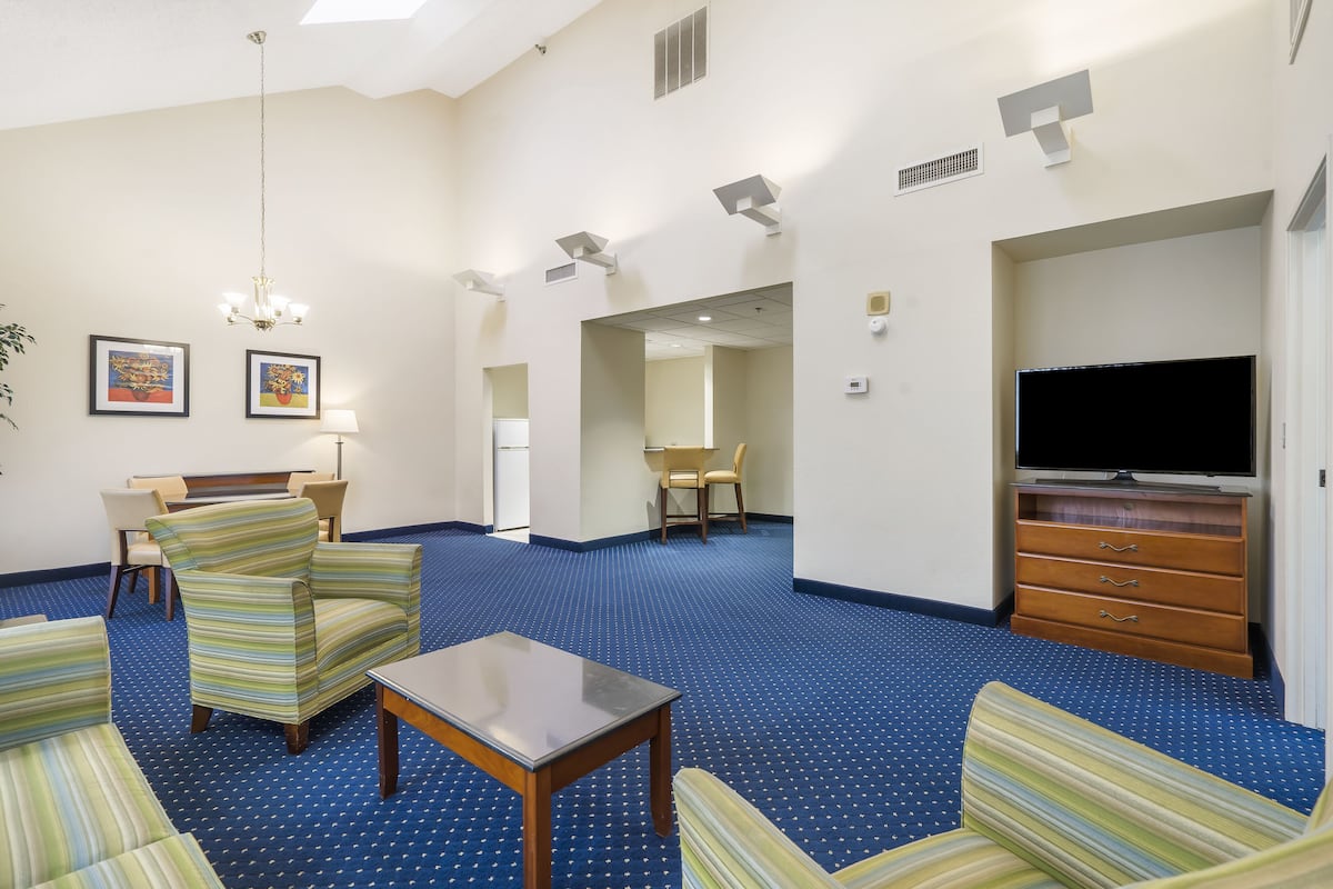 Quarto de hotel com poltronas listradas, uma mesa pequena, televisão e tapete azul.