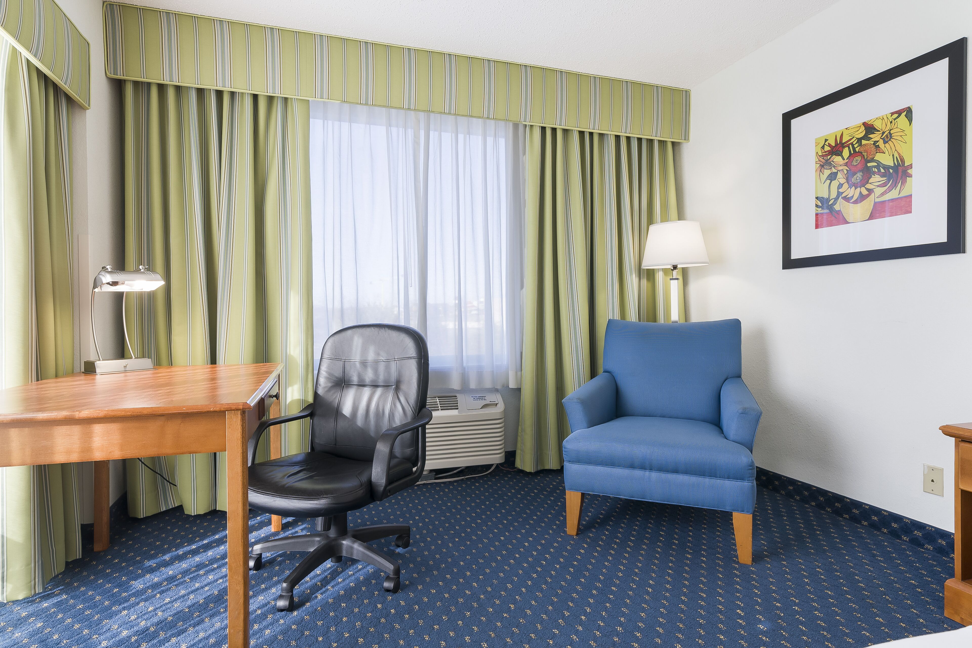 Habitación de hotel con un escritorio de madera, una silla de oficina negra, un sillón azul, una lámpara de pie y una obra de arte enmarcada en la pared.