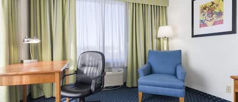 Habitación de hotel con un escritorio de madera, una silla de oficina negra, un sillón azul, una lámpara de pie y una obra de arte enmarcada en la pared.