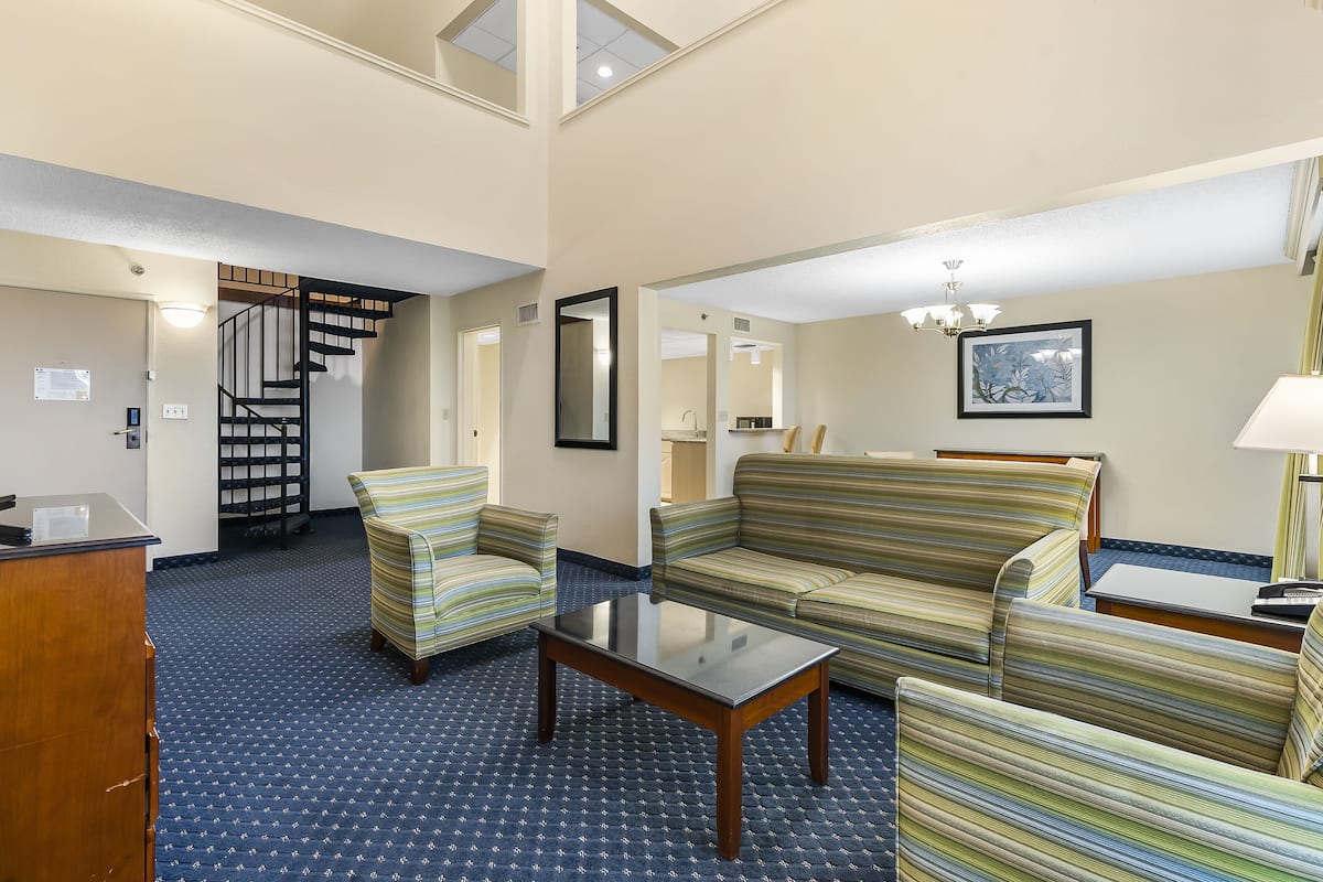 Um hall de hotel com sofás listrados, uma mesa de centro, uma escrivaninha e uma escadaria que leva a um andar superior.