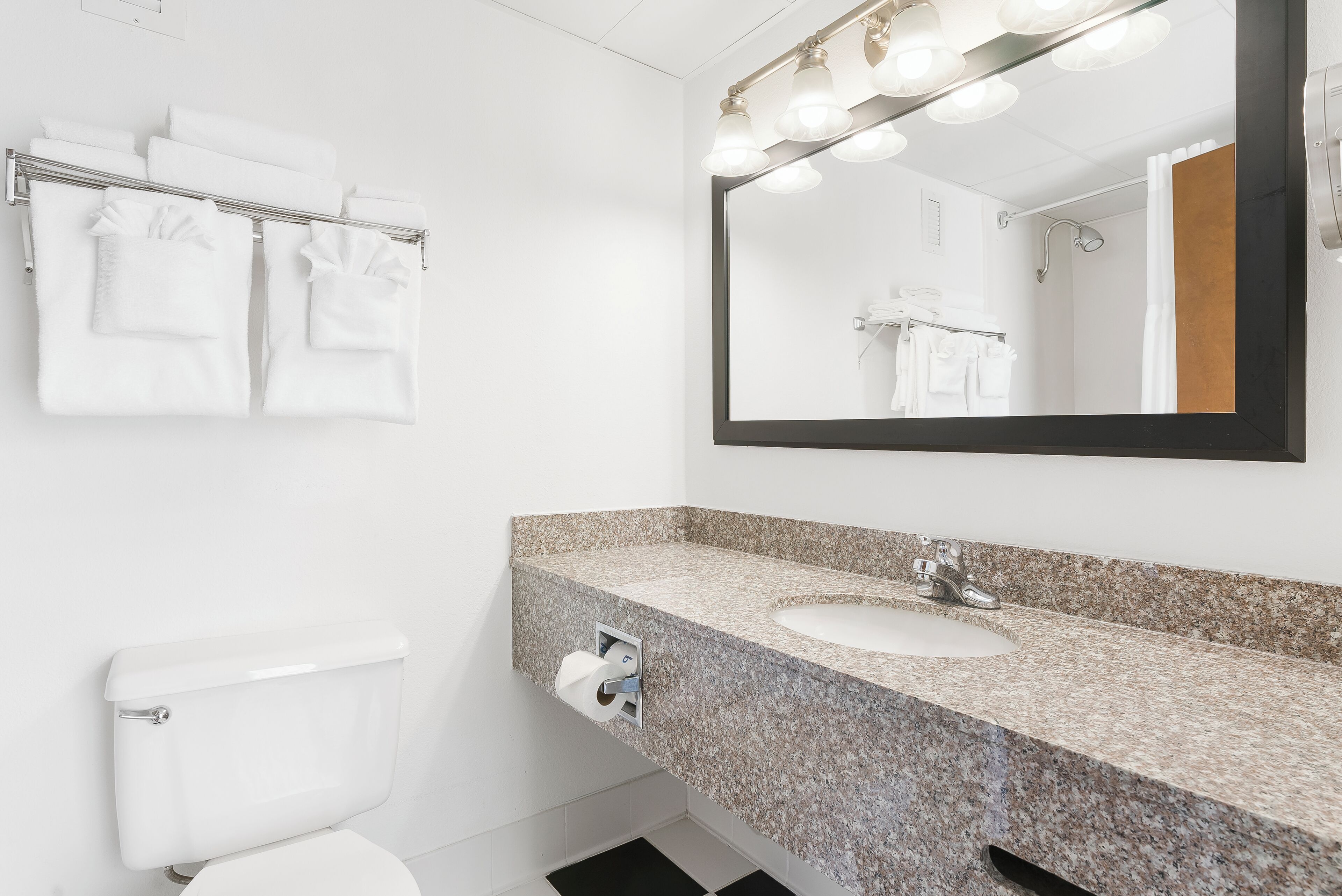 A modern bathroom with a granite countertop sink, a large mirror with lights, and a towel rack with white towels.