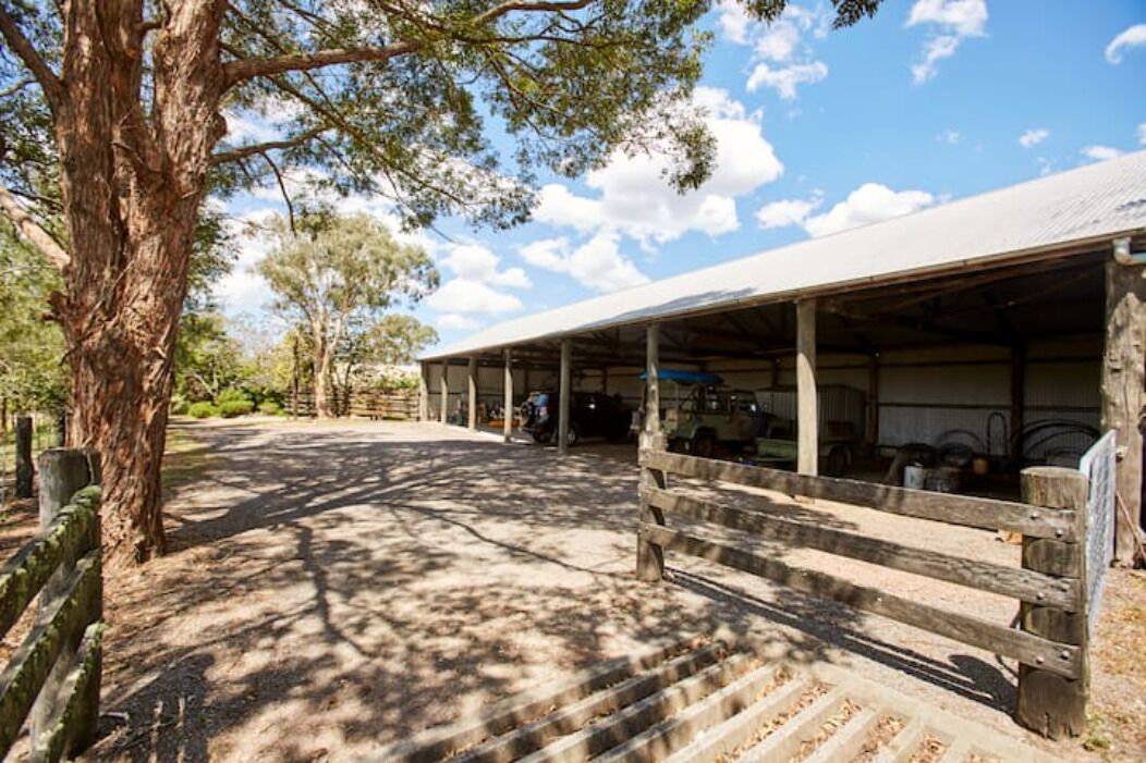 A covered outdoor area with a wooden bench, a large tree, and a building with a metal roof.