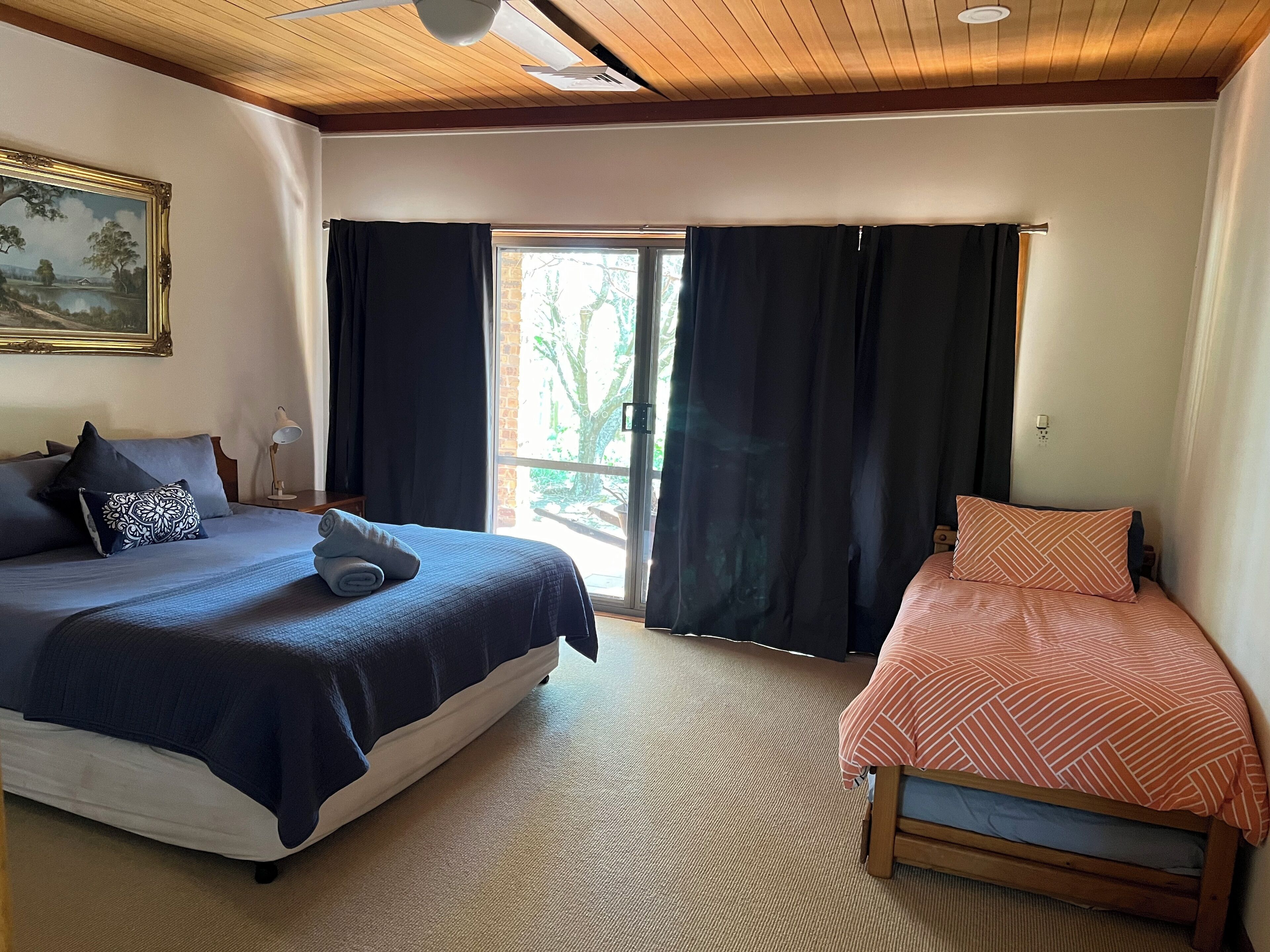 Une chambre avec un grand lit, un plus petit lit, un tableau au mur et une fenêtre avec des rideaux.