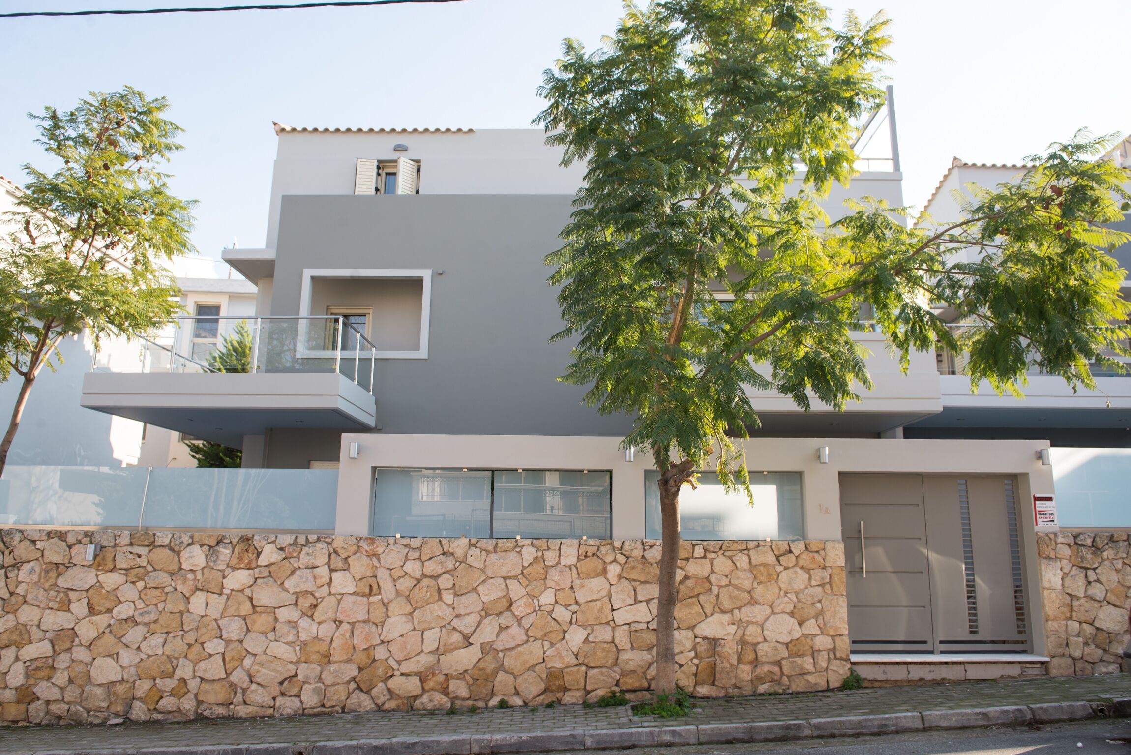 Une maison moderne à deux étages, dotée d’un mur en pierre, d’un balcon et d’un arbre devant.