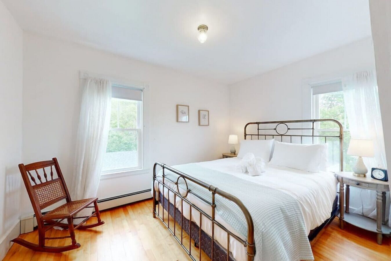 A bedroom with a metal bed frame, white bedding, a wooden rocking chair, and two bedside tables with lamps.