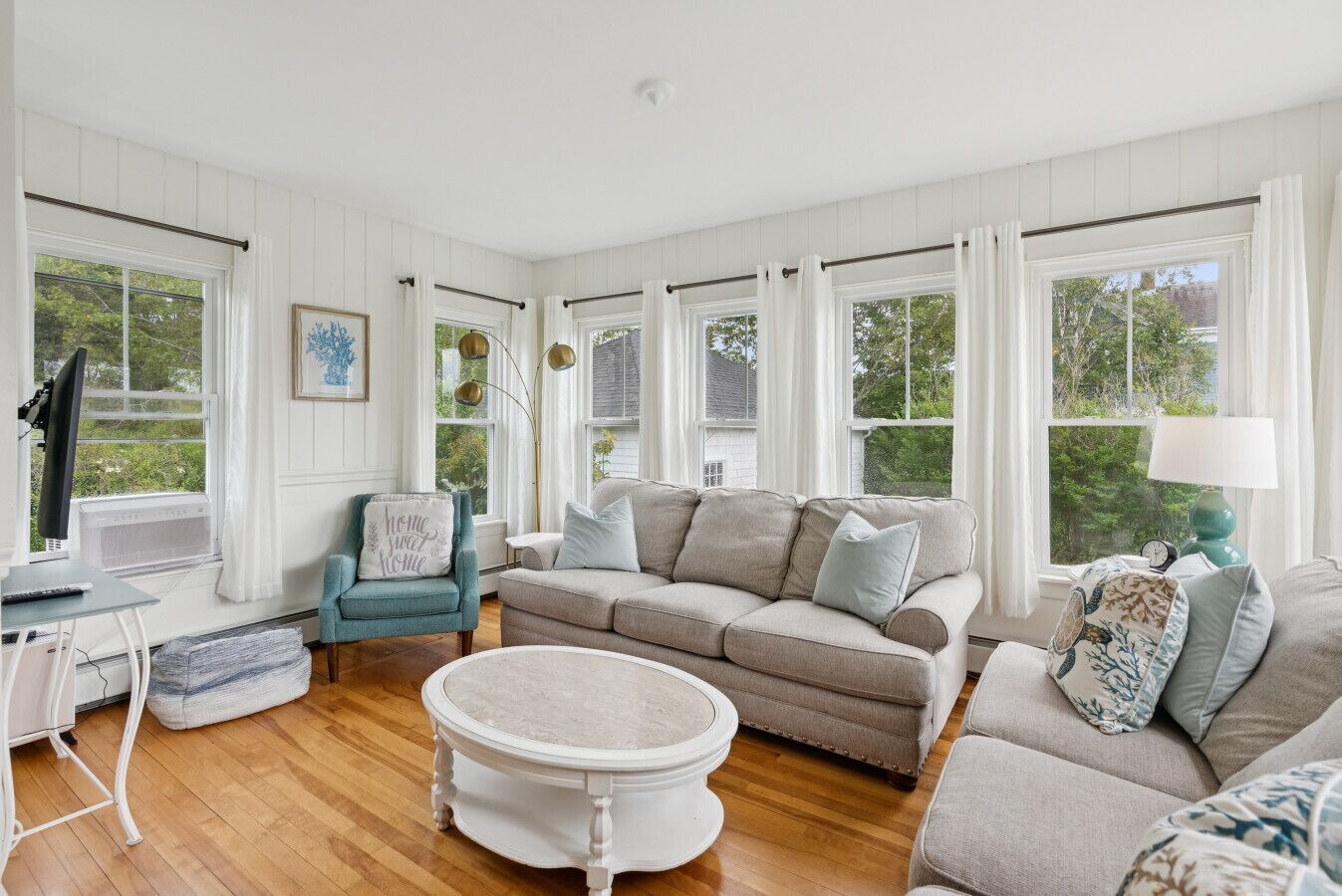 A spacious living room with a beige sofa, a teal armchair, and a round coffee table. There are large windows with white curtains, a television, and a lamp on a side table.