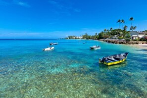 A clear tropical beach with several boats anchored in the water, a rocky shoreline, and a small building with a white roof. - Enchanting Beachfront Retreat - Leamington Cottage 1 Bedroom Villa by Blue Sky Luxury (Speightstown)
