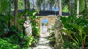 A pathway leading to a building with a view of the sea, surrounded by tropical plants and statues. - Enchanting Beachfront Retreat - Leamington Cottage 1 Bedroom Villa by Blue Sky Luxury (Speightstown)