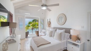 A bedroom with a bed, bedside tables, a ceiling fan, a view of palm trees, and a door leading to a balcony.