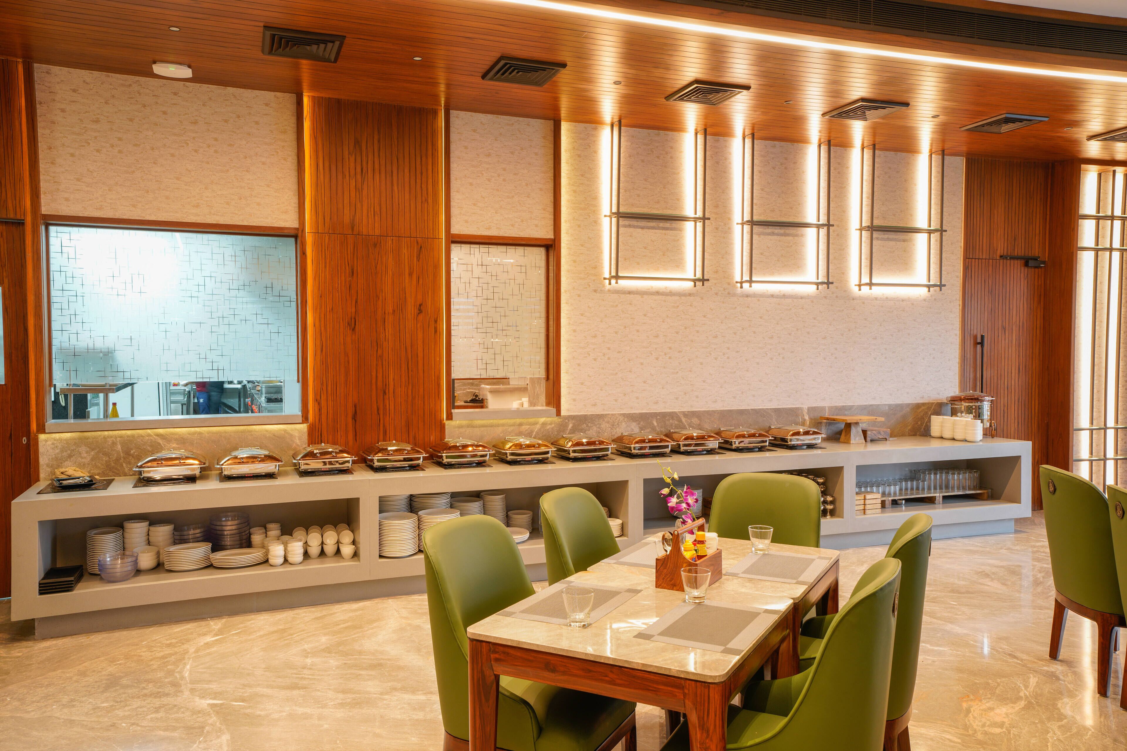 Un espace repas moderne avec un long comptoir où sont présentés différents plats, des chaises vertes et un sol en marbre.