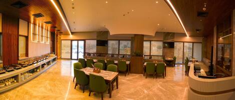A spacious hotel dining area with multiple tables and chairs, a buffet counter on the left, and large windows with blinds.
