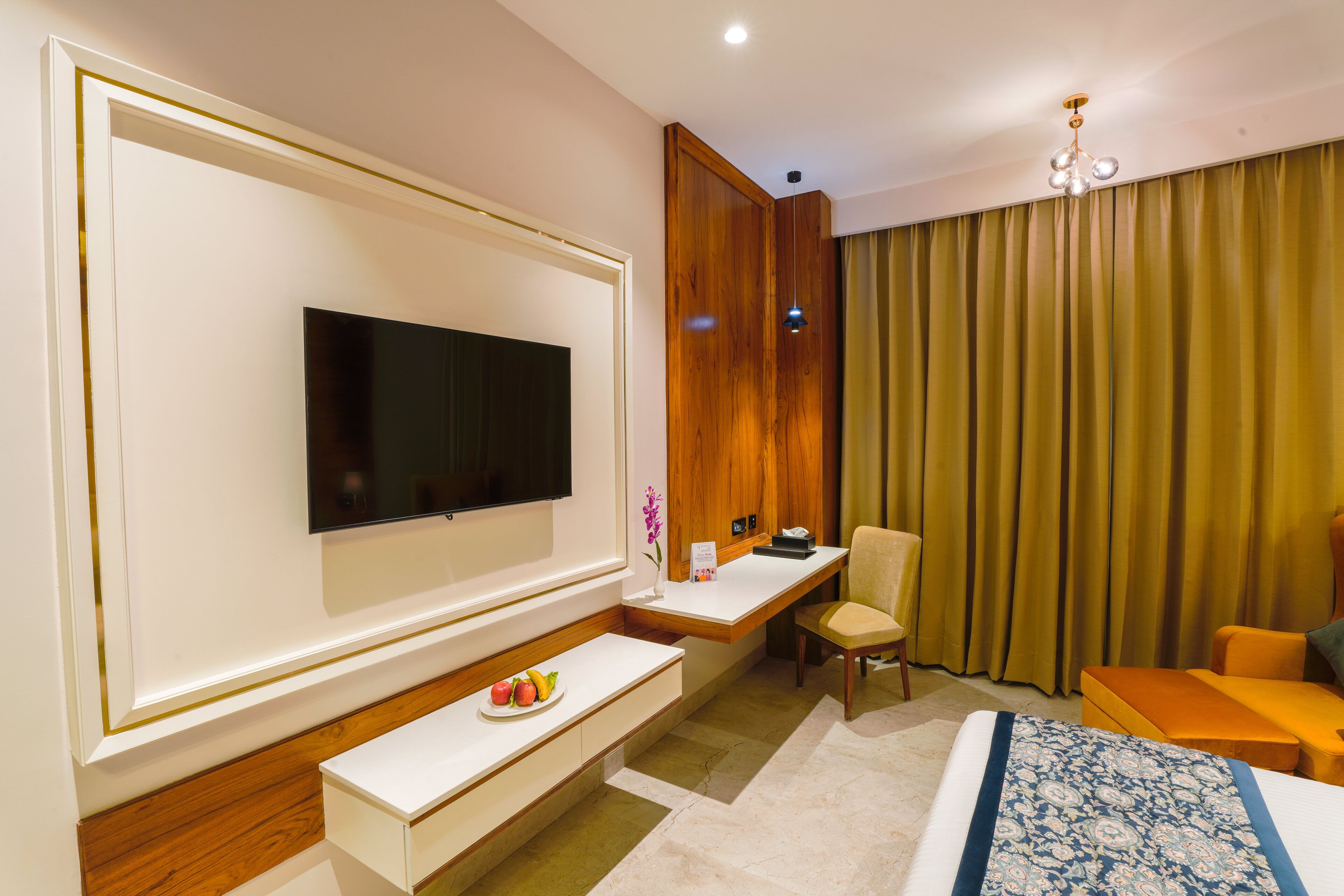 A hotel room with a flat-screen TV, a wooden desk, a comfortable chair, a sofa, and a bed with a patterned bedspread.