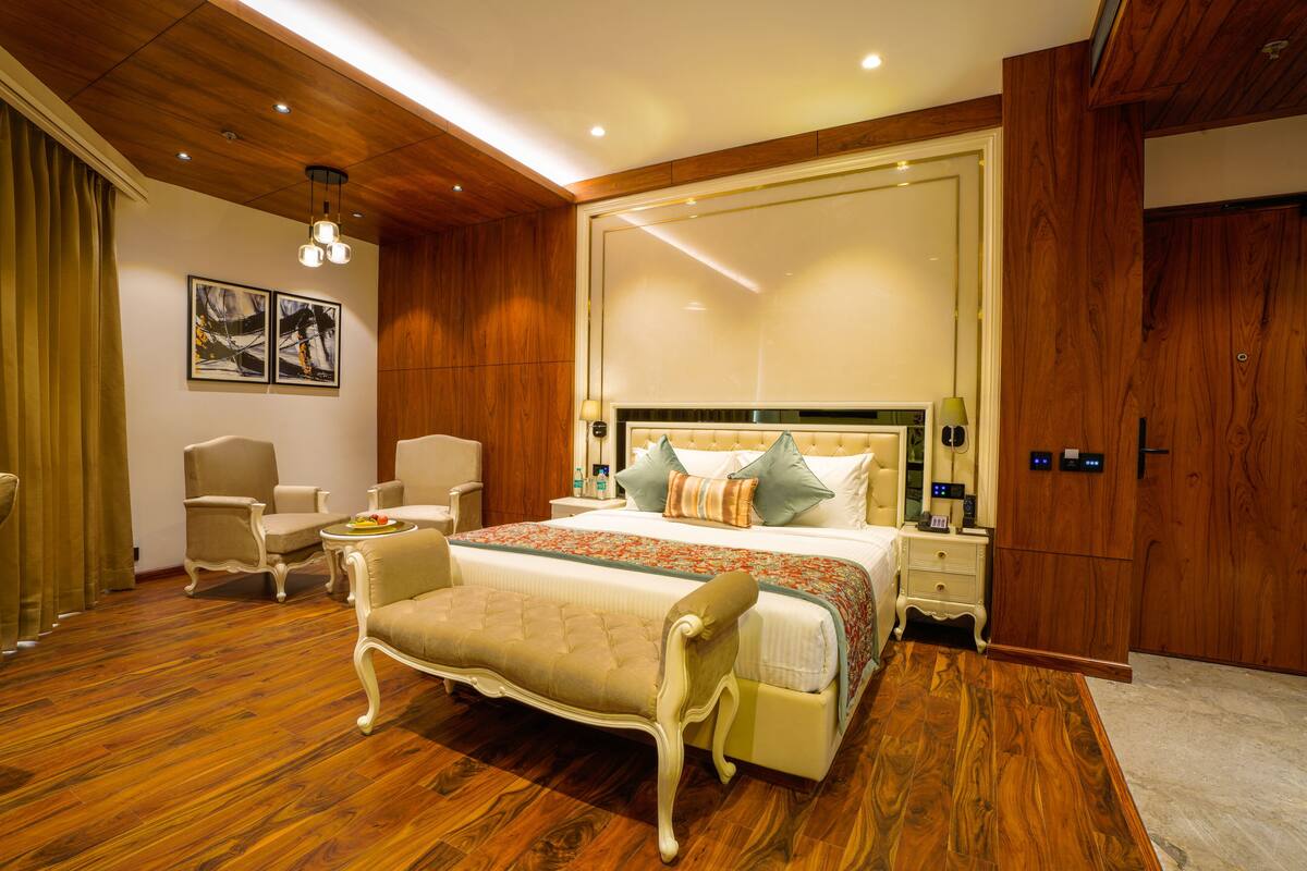 A bedroom with a large bed, a bench, two chairs, a bedside table with a lamp, and a mirror.