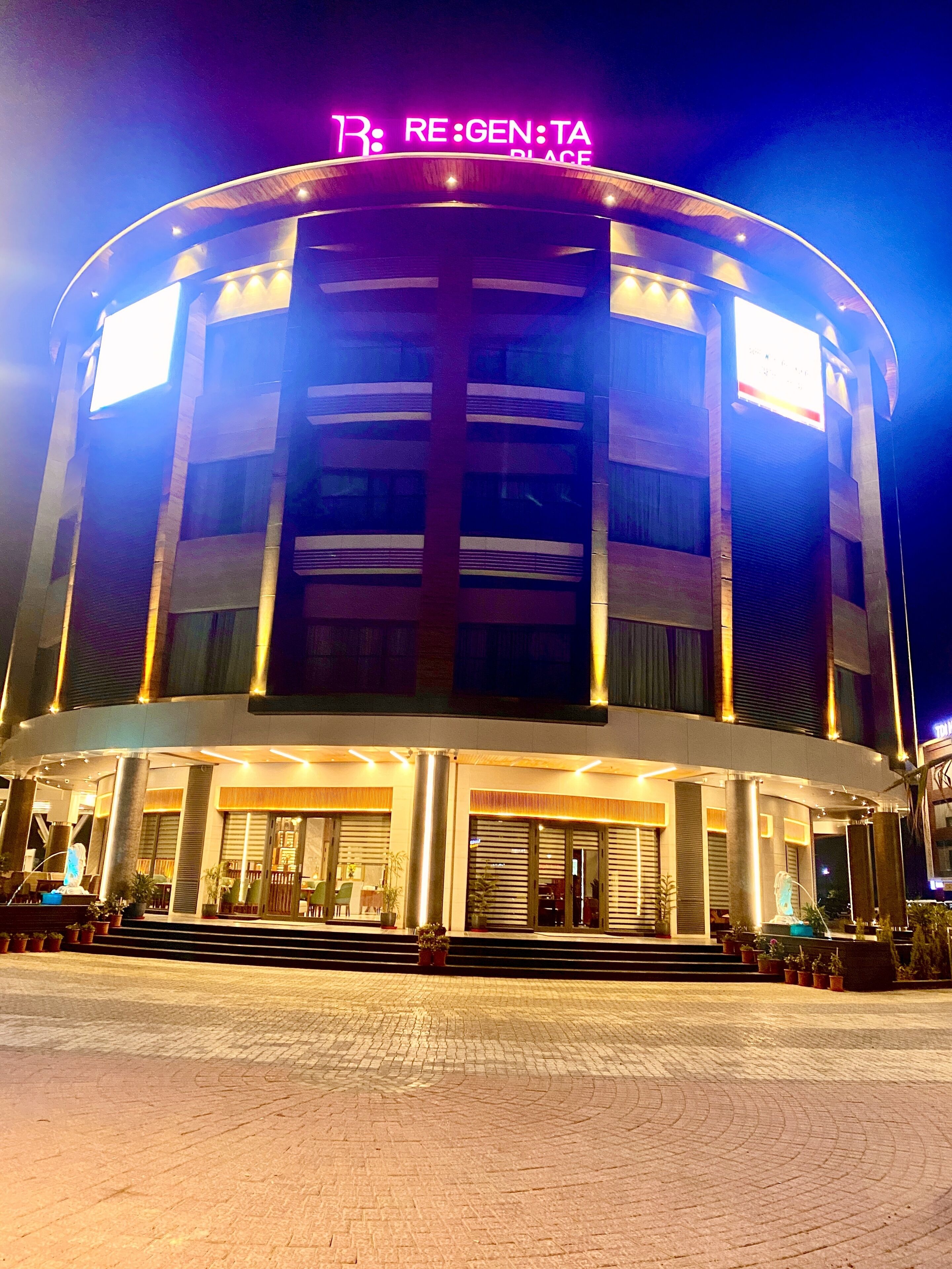 A modern hotel with a curved facade, illuminated signage, and a well-lit entrance area with potted plants.