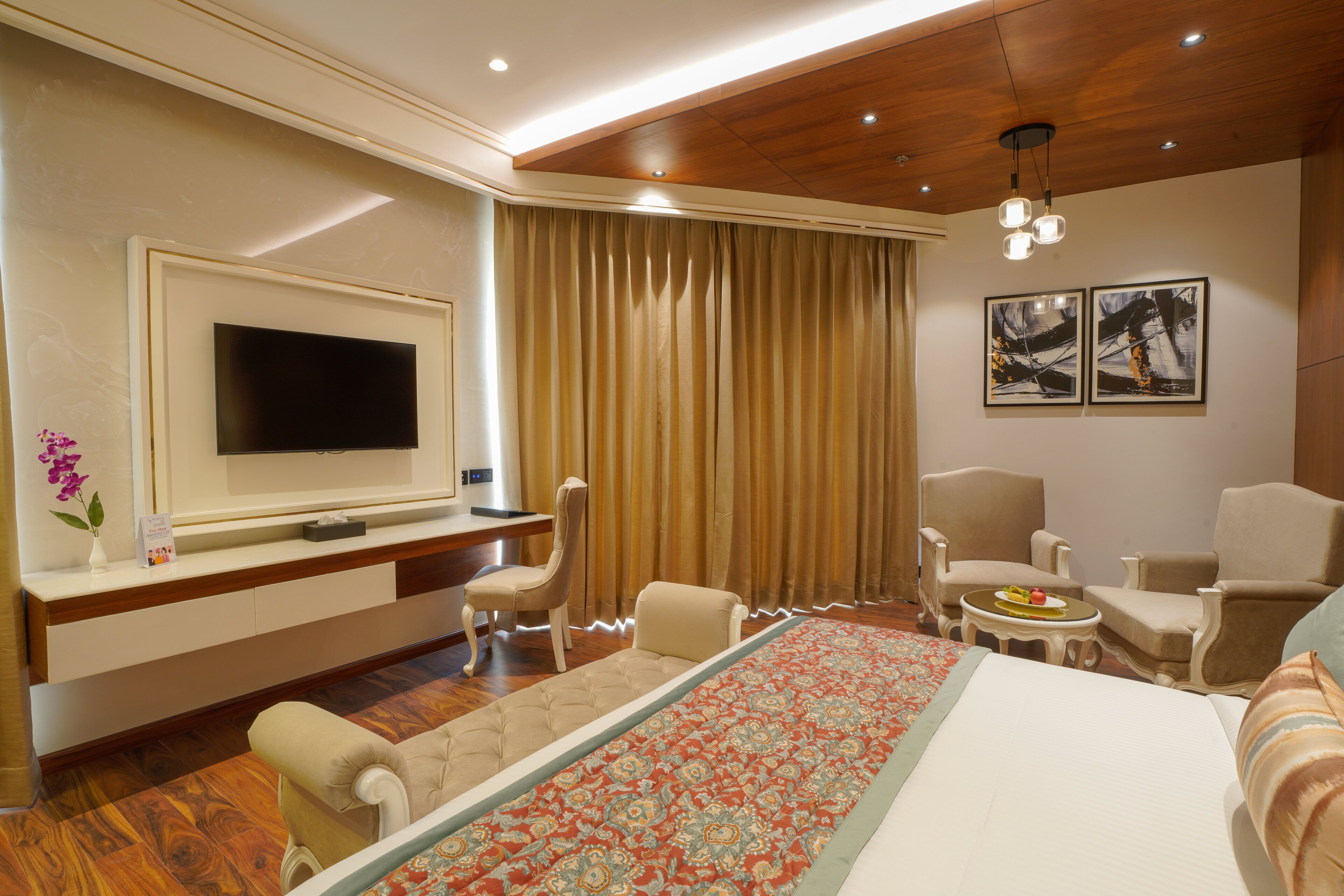 A hotel room with a large bed, a TV, two armchairs, a small table, and a large window with curtains.