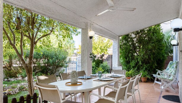 Une terrasse couverte, avec une table et des chaises, entourée de verdure et offrant une vue sur la rue.