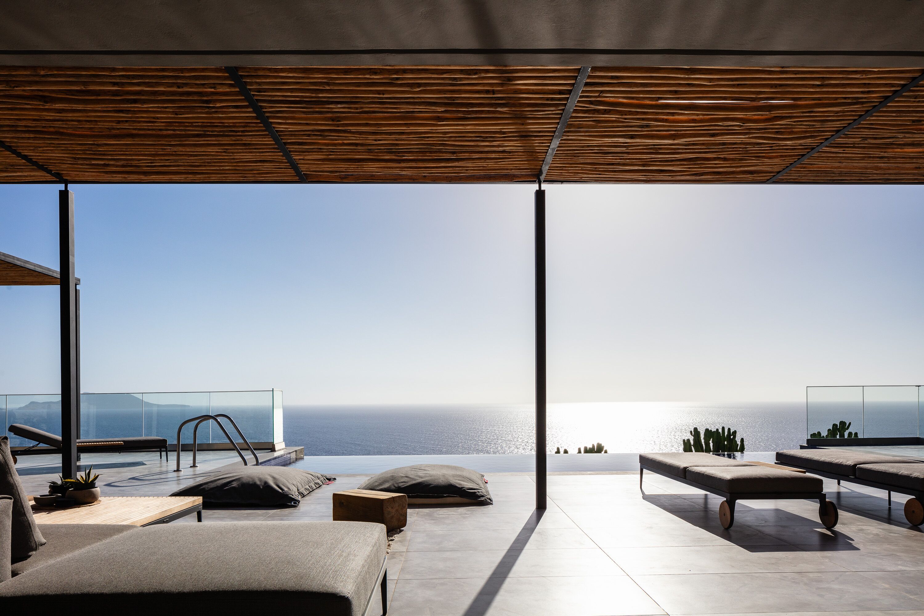 Ett modernt utomhusområde med loungekänsla och utsikt över havet, med en träpergola, bekväma loungestolar och ett glasräcke.