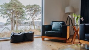 A modern living room with a leather sofa, a glass coffee table, and a view of trees and a body of water. - Carnanamish a Luxurious Cottage in Tobermory (Tobermory)