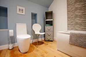 A bathroom with a toilet, a chair, a shelf unit, and a bathtub.