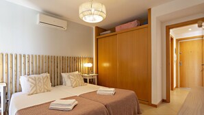 A hotel room with two beds, a wardrobe, a bedside table with a lamp, and an air conditioning unit. - Cabanas Gardens, 4px, pool viewr (Cabanas de tavira)