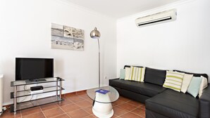 A living room with a TV on a glass stand, a black sofa with striped cushions, a floor lamp, and an air conditioner on the wall.