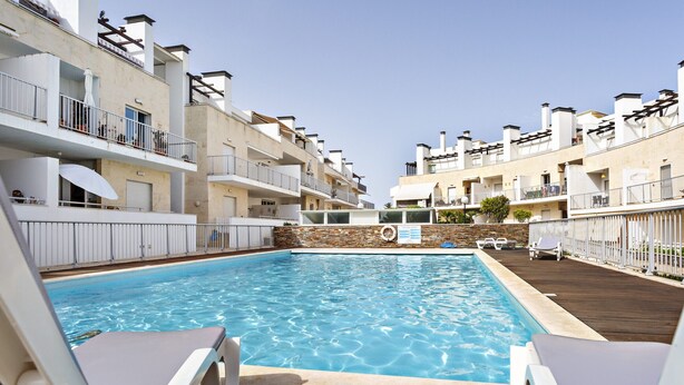 A swimming pool with lounge chairs, surrounded by multi-story buildings with balconies and a stone wall. - Cozy apartment, access to swimming pool, barbecue (Tavira)