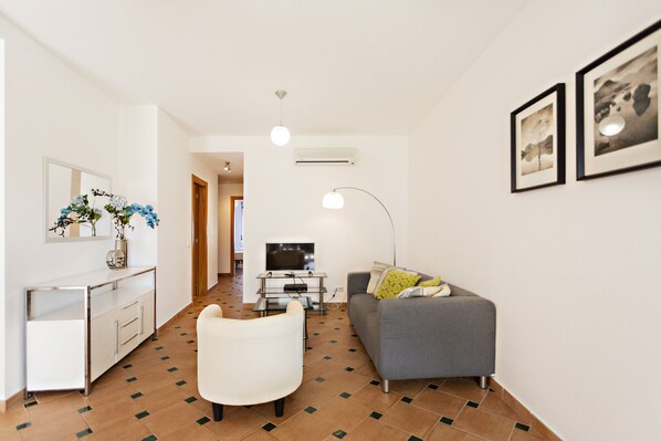 A modern living room with a sofa, a white armchair, a TV, and a desk with a vase of flowers.