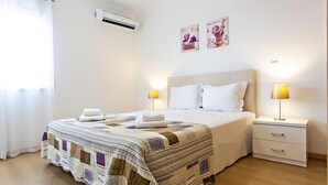 Un llit ben fet amb un edredó, dues làmpades de nit, una tauleta de nit amb calaixos i una unitat de climatització a la paret.