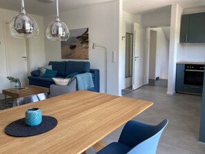 Uma sala de estar moderna com uma mesa de jantar de madeira, cadeiras azuis e um sofá com almofadas. Há também uma área de cozinha com fogão e armários.