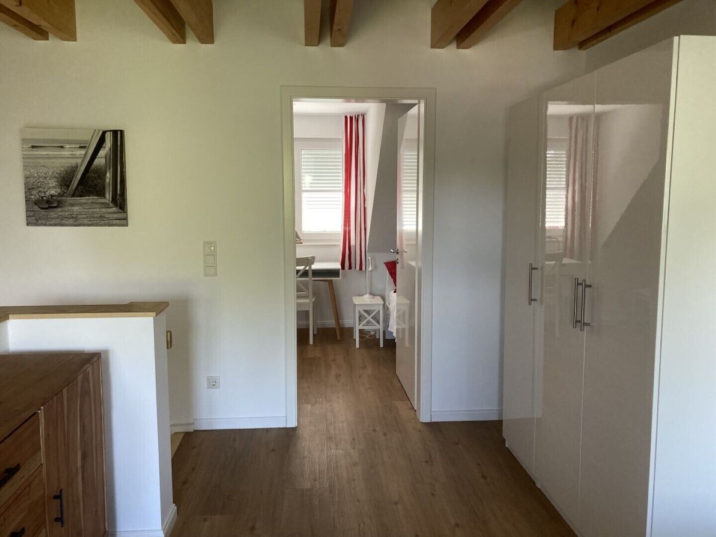 A hallway with wooden flooring, white walls, and a door leading to a room with a window and curtains.