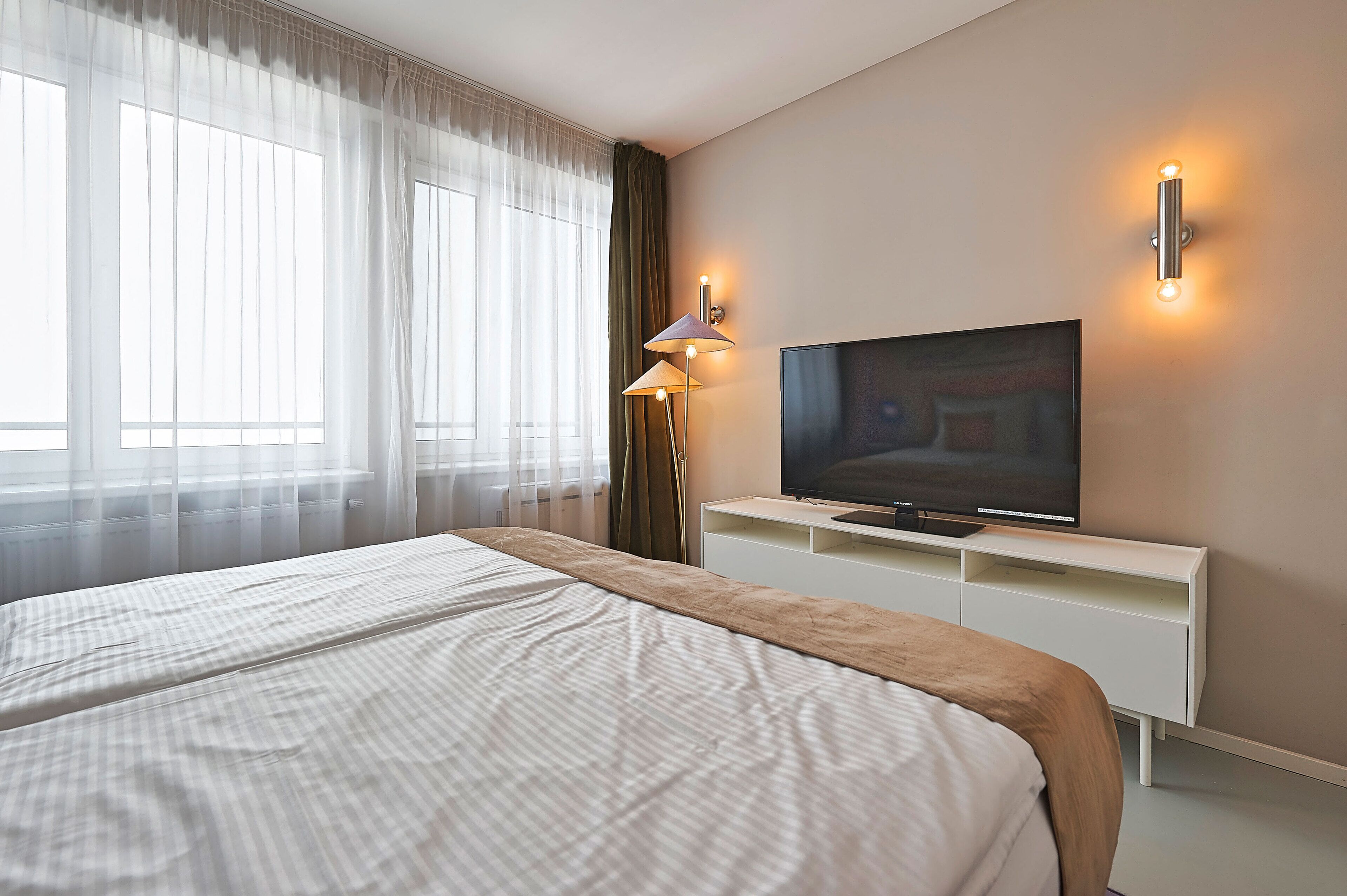 A hotel room with a large bed, a flat-screen TV on a white TV stand, a floor lamp, and sheer curtains on the windows.