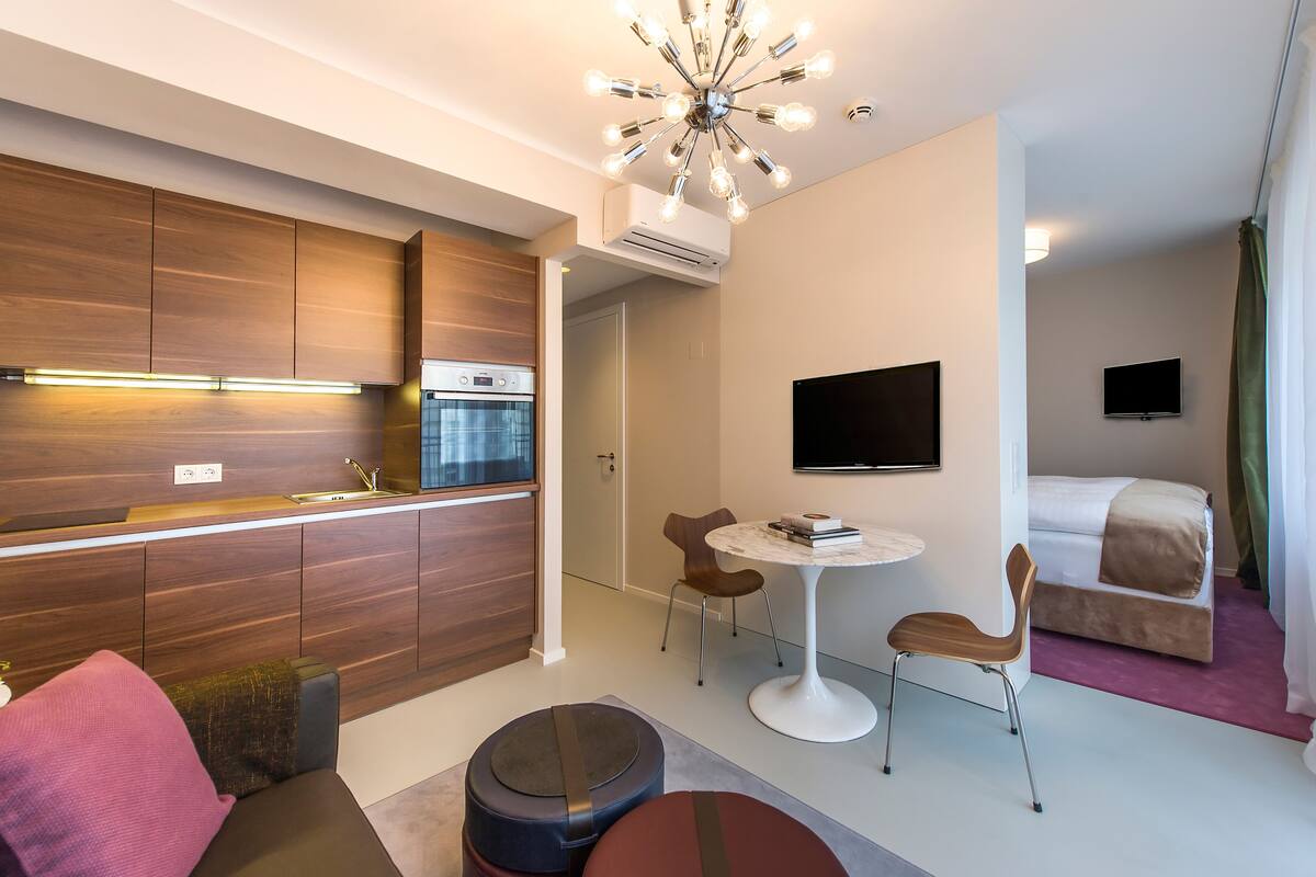 A modern hotel room with a kitchenette, a dining area, a bed, and a TV.
