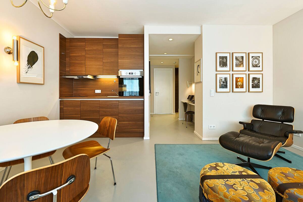 A modern kitchen with wooden cabinets and a built-in oven, a dining area with a white table and wooden chairs, and a living area with a black leather sofa and ottomans.