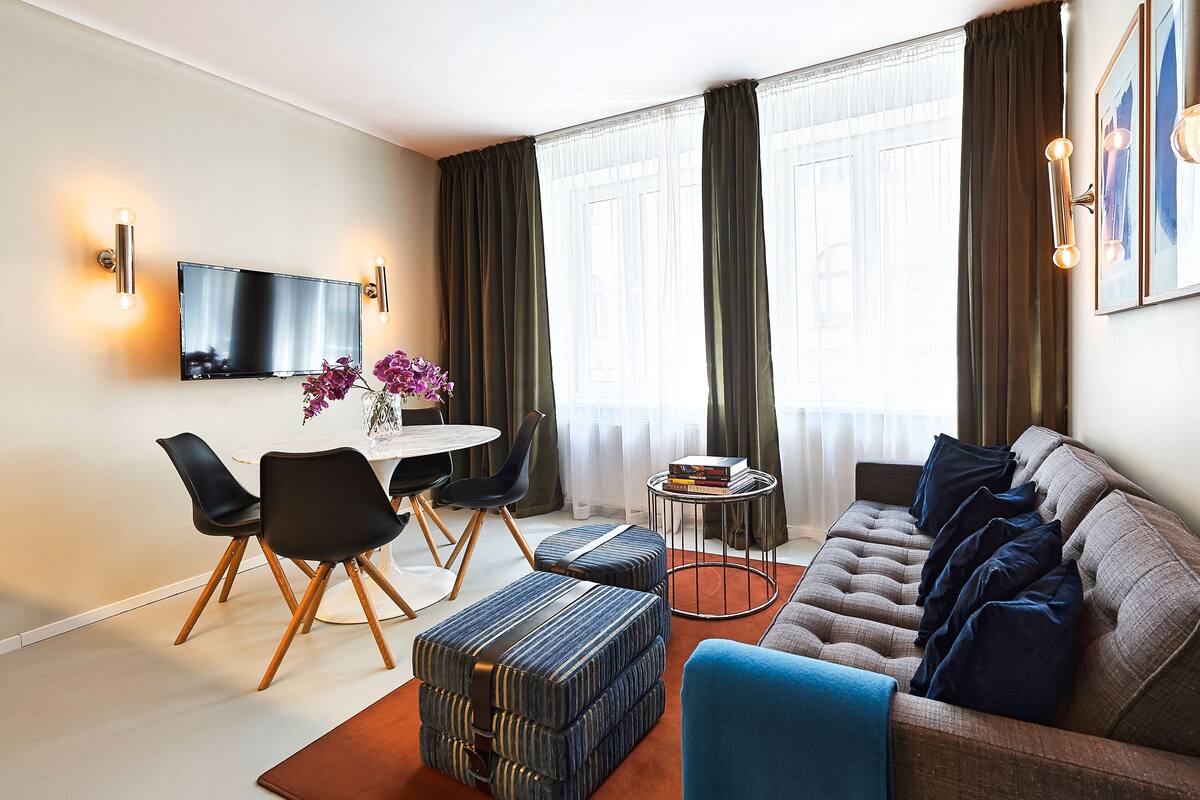 A modern living room with a sofa, ottoman, dining table, chairs, and a TV mounted on the wall.