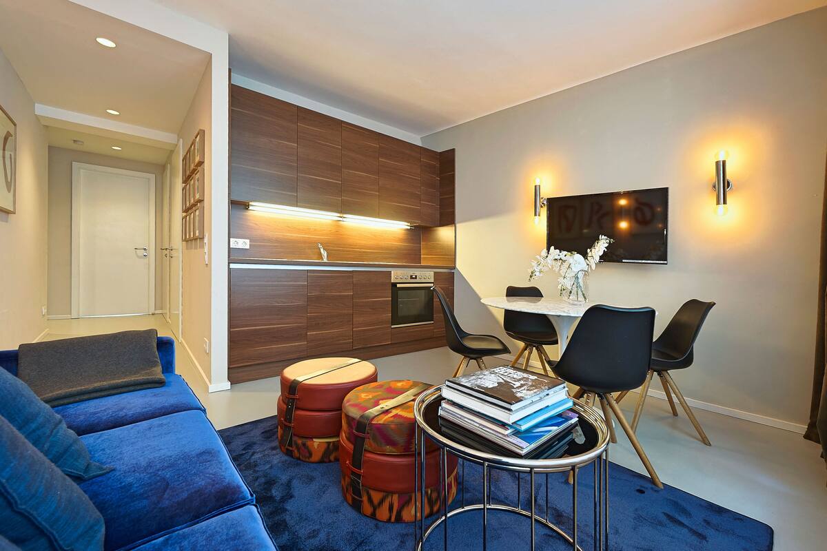 A modern living room with a blue sofa, a small round table with books, and a dining area with chairs and a TV.
